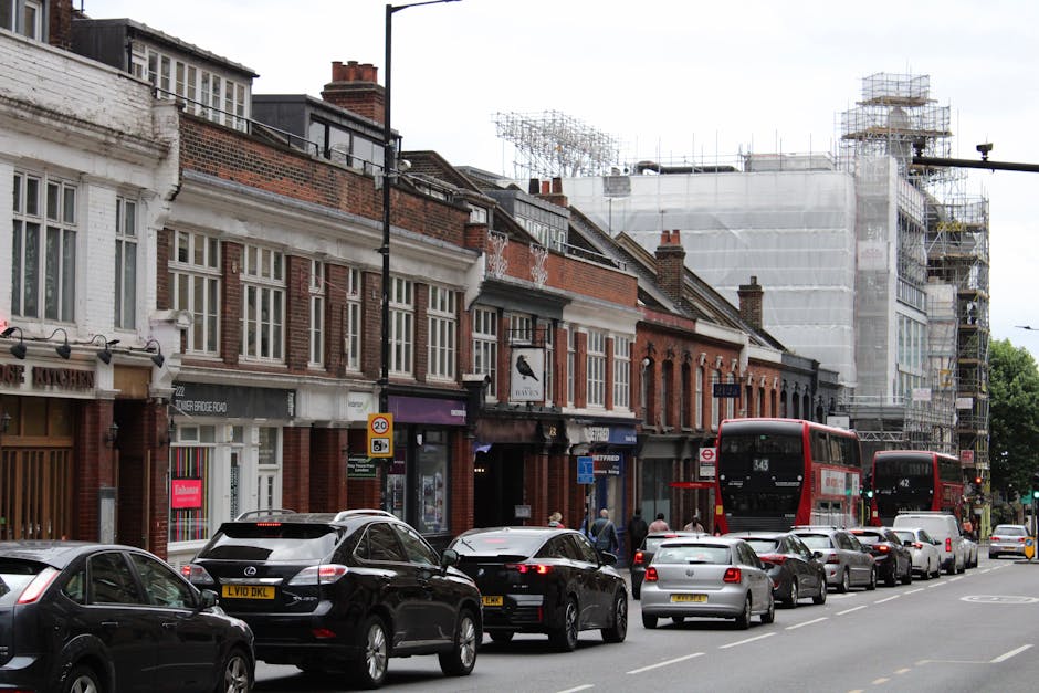 A busy street scene in an urban area featuring a row of traditional brick terraced buildings with white window frames and decorative brickwork, some of which are under construction or renovation, as indicated by scaffolding and protective sheeting covering parts of taller structures. The street is lined with parked cars, including a black hatchback, a silver sedan, and a dark SUV, with a line of additional vehicles, including a white van and multiple smaller cars, extending into the background. Two red double-decker buses, numbered 343 and 42, are navigating the street amid pedestrian activity on the sidewalk, where a few people are walking or waiting. The environment appears overcast with a cloudy sky, and the street scene showcases typical urban traffic and building maintenance, relevant to private waste management and local rubbish removal services such as those offered by Rubbish Clearance Harrow, especially in contexts involving street-access limitations and on-site clearance for property renovations.