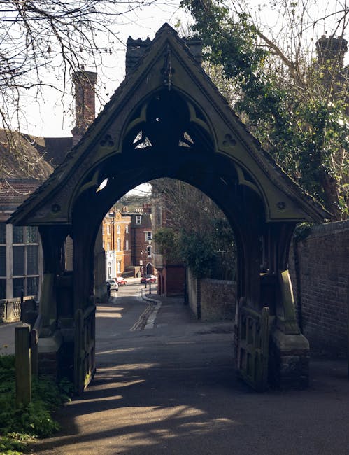 A traditional black wooden gate with intricate Gothic-style arch detailing forms the entrance to a narrow street in a residential area. The gate's textured surface shows weathered wood and decorative carved elements, with the pointed arch rising prominently at the top. Behind the gate, a small alleyway extends, paved with tarmac and lined by brick walls on either side, leading to a street with parked cars and mid-19th-century brick buildings visible in the background. On the left, a portion of a brick building with large windows and a chimney can be seen, while the right side features a brick wall and some overhanging tree branches partially covered with leaves. The scene is illuminated by natural sunlight, casting shadows on the ground and highlighting the detailed craftsmanship of the gate, which acts as an architectural feature and potential access point in the context of private waste handling or on-site clearance, as offered by services such as Rubbish Clearance Harrow.
