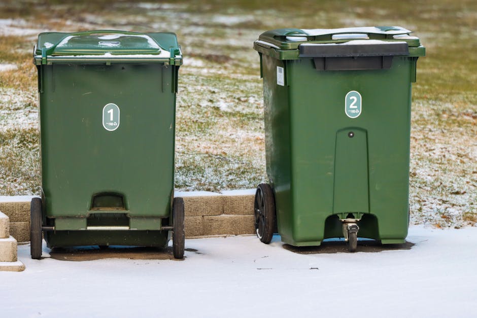 Two green wheeled rubbish bins are positioned side by side on a pavement in an outdoor area with a low brick wall behind them. The bin on the left has a flat, closed lid and a label with the number 1, while the bin on the right features a slightly domed lid with a label marked with the number 2. Both bins are made of durable plastic with a smooth finish, and the wheels are black with a textured surface for movement. The ground beneath the bins shows patches of snow and ice, indicating cold weather, while the backdrop includes a grassy area with a dusting of snow and a slightly blurred environment. The bins are in good condition, with some minor scratches, and are intended for general waste or garden refuse collection, reflecting private waste disposal options potentially available through services like Rubbish Clearance Harrow. The professional scene emphasizes on-site waste management in a residential or public outdoor setting, aligning with alternative rubbish removal methods outside of local authority collections.