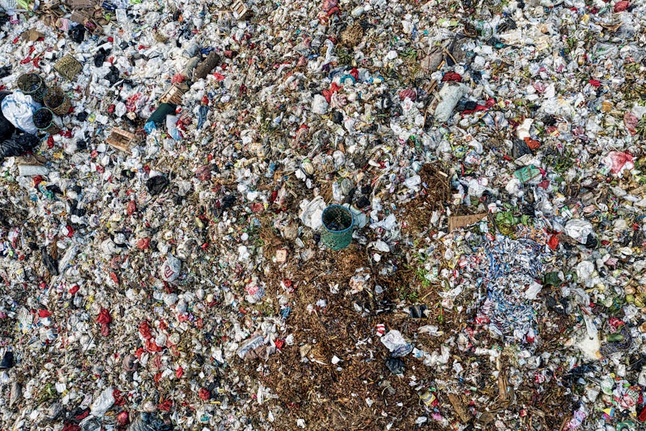 An expansive aerial view of a large landfill site filled with various types of waste. The surface is densely covered with scattered plastic bottle caps, wrappers, paper waste, and decomposing organic matter. Patches of soil are visible where waste has been disturbed or piled up, with some areas showing darker, wetter layers of decomposing material. Among the debris, there are small fragments of cardboard and plastic containers, with some items partially buried or overlapping others. The scene includes a few scattered green plants emerging from the waste, indicating incomplete decomposition. The overall environment appears cluttered and unmanaged, highlighting the scale and mess of waste accumulation. This image underscores the importance of proper rubbish disposal and waste management, which companies such as Rubbish Clearance Harrow facilitate through alternative rubbish collection and on-site clearance services for clean and sustainable waste handling, especially considering local council rules for garden and household waste disposal.
