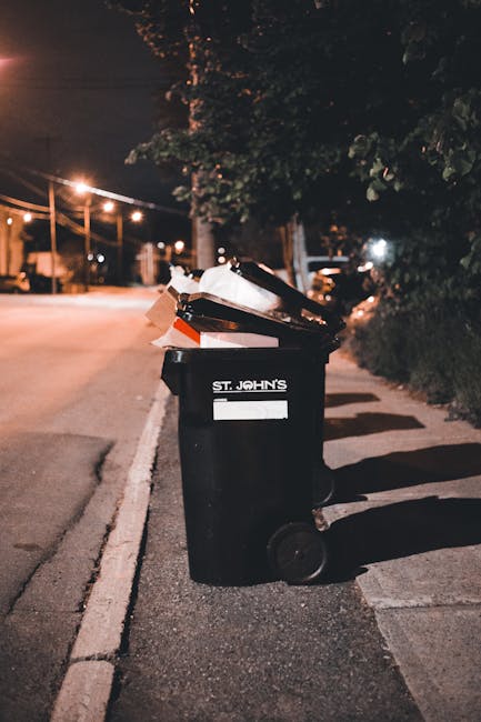 Two green wheeled rubbish bins are positioned side by side on a pavement in an outdoor area with a low brick wall behind them. The bin on the left has a flat, closed lid and a label with the number 1, while the bin on the right features a slightly domed lid with a label marked with the number 2. Both bins are made of durable plastic with a smooth finish, and the wheels are black with a textured surface for movement. The ground beneath the bins shows patches of snow and ice, indicating cold weather, while the backdrop includes a grassy area with a dusting of snow and a slightly blurred environment. The bins are in good condition, with some minor scratches, and are intended for general waste or garden refuse collection, reflecting private waste disposal options potentially available through services like Rubbish Clearance Harrow. The professional scene emphasizes on-site waste management in a residential or public outdoor setting, aligning with alternative rubbish removal methods outside of local authority collections.