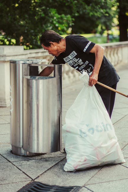 A middle-aged man with short, dark hair is seen wearing a black t-shirt with white text and graphics. He is outdoors on a paved walkway, near a stone balustrade with lush green trees in the background. The man is using his right hand to place a large, white plastic bag filled with rubbish into a modern, cylindrical metal waste bin designed for waste separation, featuring multiple compartments. He holds a wooden stick or pole in his left hand to assist with handling the bag. The scene appears to be during daylight with natural light illuminating the area, emphasizing the polished, reflective surface of the waste bin and the textures of the man's clothing and the paved ground, which is typical of public space cleaning or waste management activity, aligning with the concept of alternative waste handling and rubbish removal services or on-site clearance by private providers like Rubbish Clearance Harrow.