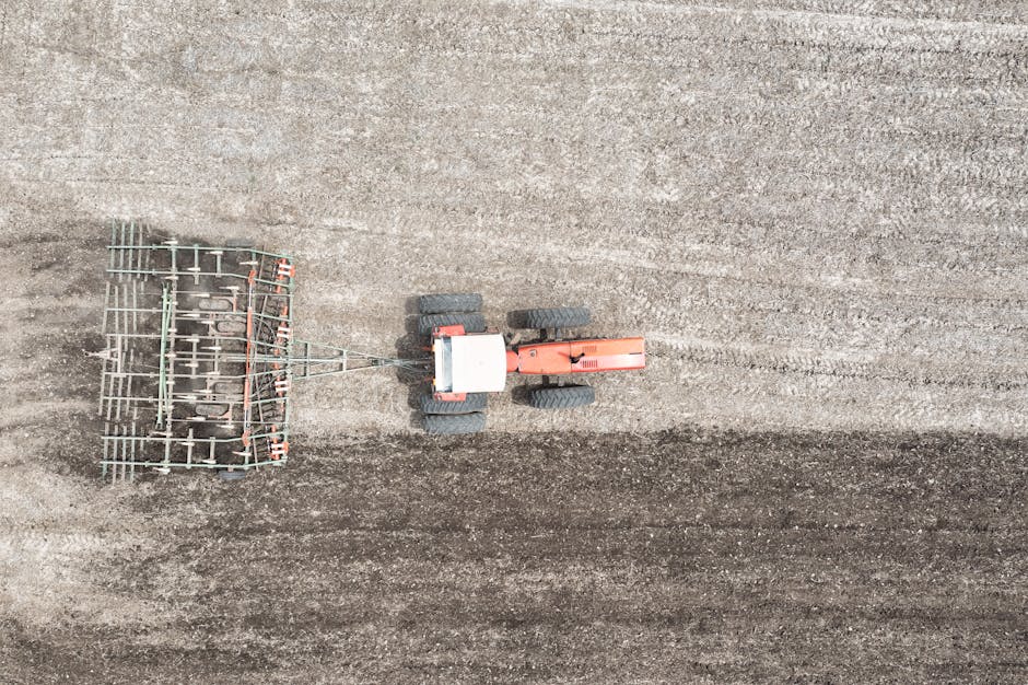 An aerial view of a small orange and grey tractor with four large black rubber tires operating on a tilled, earthy brown soil surface, pulling a metal harrow attachment with multiple metal tines arranged in a grid pattern. The soil shows distinct parallel furrows created by previous cultivation, and the scene is set outdoors under natural daylight, with no other objects or structures visible. The image emphasizes machinery used in agricultural or land preparation activities, similar to the equipment employed in on-site rubbish or waste clearance services offered by Rubbish Clearance Harrow, which manage varied waste removal tasks in settings such as gardens or construction sites.