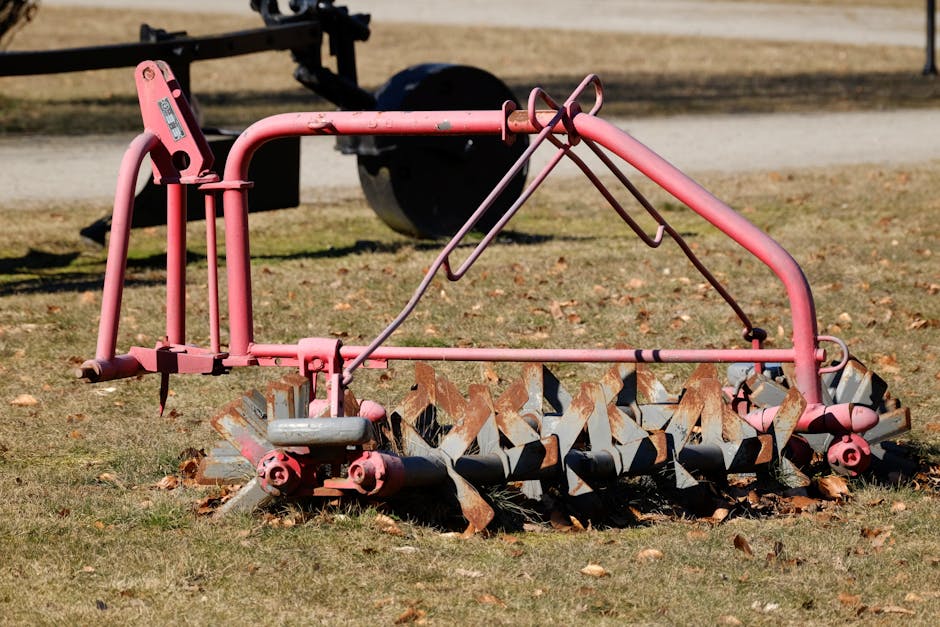 A weathered pink lawn tractor attachment with a metal frame and large, rusted, spiked blades at the base is situated on a patch of grass, surrounded by fallen leaves. The attachment features a curved handle and support bars, with some paint chipping exposing the underlying metal. The blades are chipped and rusty, showing signs of frequent use, and are partially embedded in the soil, with leaves caught within the blades. The environment appears to be an outdoor area, possibly a garden or park, with a gravel path or driveway visible in the background along with a black piece of equipment or trailer tire, indicating proximity to agricultural or gardening activities. The scene is lit by natural daylight, casting shadows on the grass and highlighting the textures and colors of the rusty blades and worn metal frame, conveying a sense of outdoor maintenance equipment likely slated for disposal or recycling as part of a waste removal service by Rubbish Clearance Harrow.