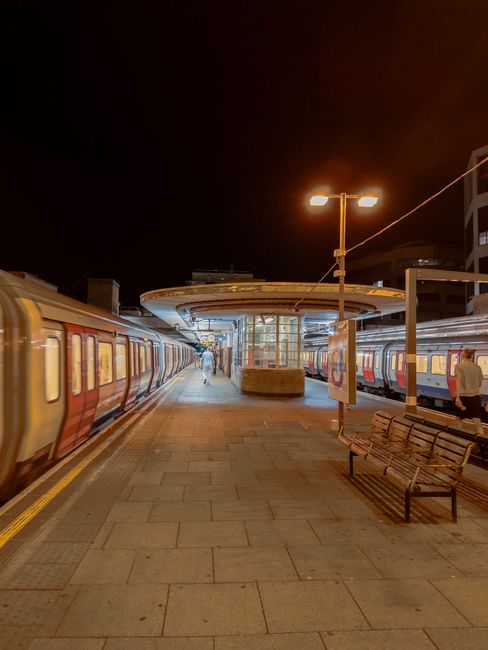 A nighttime scene at a train station platform shows two trains parked on either side of the paved walkway. The trains have a two-tone color scheme, with silver and red or blue sections, and windows along their sides. The platform is illuminated by tall street lamps casting a warm yellow glow, and there are a few passengers visible, some walking towards the station building in the background, which features glass walls and a flat, circular roof. In the foreground, there is a park bench and a small, round planter or decorative structure near the station's entrance. The surrounding environment includes nearby buildings with multiple stories, suggesting an urban setting. The scene captures a calm, quiet moment during the night, illustrating an element of private or independent travel that may relate to the themes of alternative transport or private waste disposal, aligning with the topics of rubbish removal and waste management services provided by Rubbish Clearance Harrow.