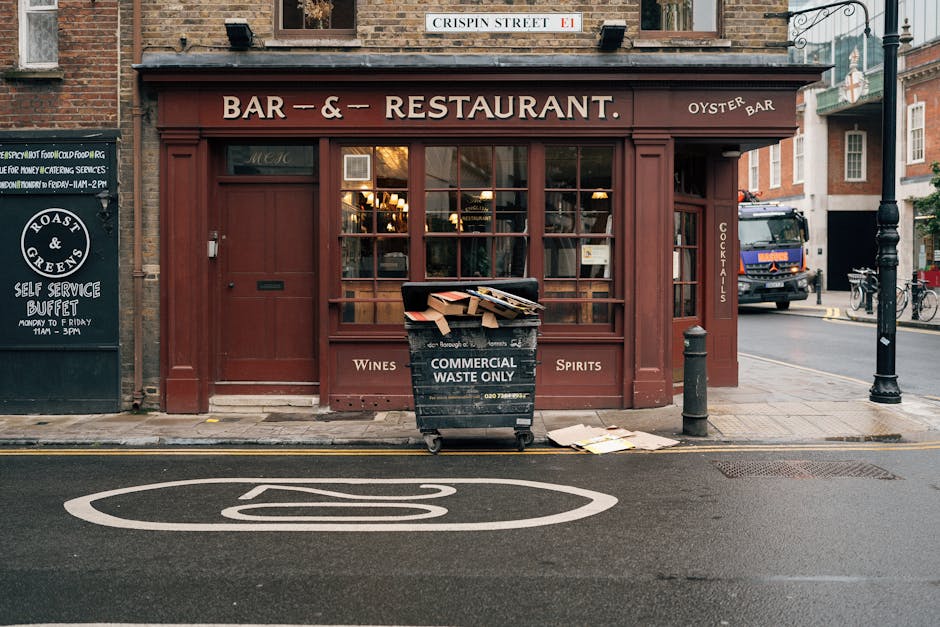 A black wheeled waste collection bin labeled 'Commercial Waste Only' positioned on the sidewalk in front of a café or bar establishment with a red-brown wooden facade. The bin is filled with flattened cardboard boxes and other waste materials. Surrounding the bin, there are small papers or cardboard pieces on the pavement. The storefront features large glass windows divided into smaller panes, with a partially visible sign reading 'Wines' and 'Spirits' on the window frame. To the left, there is a blackboard-style sign advertising a self-service buffet, and on the right side, a street lamp and a bollard are present. In the background, a parked truck and a bicycle are visible, indicating an urban street scene. The overall environment suggests an area where independent waste management, such as private rubbish collection, takes place outside commercial premises, aligning with the context of non-local authority waste disposal options supported by services like Rubbish Clearance Harrow.