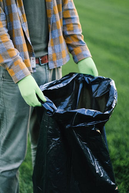 A person wearing a blue and yellow checked shirt, grey trousers, and bright green gloves is holding open a large black rubbish bag made of plastic, positioned on a grassy outdoor area. The bag is partially filled with waste and appears ready for collection or disposal, reflecting typical activities associated with rubbish removal services. The surrounding environment features a well-maintained lawn or field, with the background showing a soft-focus green landscape. The lighting is natural, suggesting daytime, and the scene emphasizes on the manual handling of waste as part of private rubbish clearance or alternative waste disposal methods, with no visible vehicles or equipment. The person's attire and the setup imply an on-site waste collection or clean-up, typical of independent rubbish removal activities managed by companies such as Rubbish Clearance Harrow.