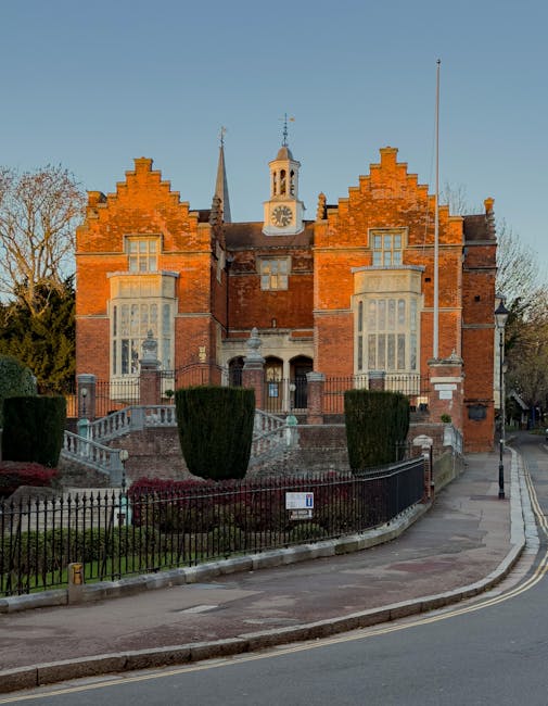 A historic brick building with an ornate architectural style, featuring a symmetrical façade with large bay windows, decorative stone accents, and a central clock tower topped with a small spire. The structure is set slightly uphill, accessible via stone steps with wrought iron railings, leading from a paved walkway bordered by a black metal fence. In front of the building, there is a small landscaped garden with neatly trimmed, rounded bushes and a patch of grass. The scene is illuminated by natural daylight, with a clear blue sky overhead, and the surrounding environment includes a curved road with a street lamp on the right side. The building appears well-maintained, showcasing a traditional design typical of historic municipal or institutional buildings, which may be an example of private or alternative premises where rubbish removal services could be relevant for maintenance or clearance purposes, as provided by Rubbish Clearance Harrow.