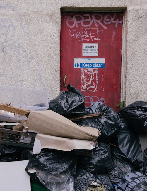 A large pile of black garbage bags, some torn open revealing waste inside, and flattened cardboard boxes are stacked in front of a red metal door with multiple graffiti tags. The door, set into a textured, off-white stucco wall, features signs including one stating 'No Money' and another with a blue and white message reading 'Keep clear.' The scene is outdoors, likely on a pavement or alleyway, with small patches of greenery near the lower right corner. The lighting is natural and evenly diffused, highlighting the disorganized rubbish that appears ready for disposal or collection by a private waste removal service. Overfilled and leaning, the waste suggests an unscheduled or independent rubbish clearance, aligning with alternative waste handling methods often employed when traditional local authority collection might not be suitable or available. Overall, the scene exemplifies typical waste accumulation in an urban environment requiring professional rubbish clearance services such as those offered by Rubbish Clearance Harrow, providing a clear visual of waste awaiting removal in an external setting, close to a service access point.