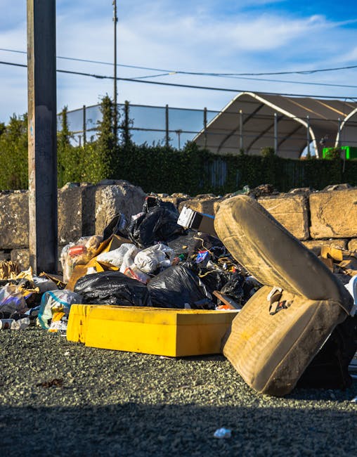 A close-up view of a roadside rubbish pile containing various waste materials, including black and clear plastic garbage bags, discarded packaging, and miscellaneous debris. In the foreground, a large, worn, beige car tire with a textured sidewall rests on the ground, partially leaning against the pile. Nearby, a bright yellow plastic container or tray lies on the gravel surface, which is uneven and coarse. The background features a stone wall made of irregularly shaped, rough-textured stones in light brown and gray tones, and behind this wall, there is a dark green hedge and some trees. Further back, a chain-link fence separates the area from a sports court or outdoor facility, identifiable by a covered structure with a curved, white, semi-transparent roof. Overhead, a clear blue sky with a few wispy clouds; several power lines run horizontally across the scene, adding an urban context. The scene is lit by natural daylight, casting shadows and highlighting the textures of the waste and environment. The image captures a typical example of unmanaged roadside waste, possibly related to private rubbish clearance services in the Harrow area, illustrating the importance of professional rubbish removal options like those provided by Rubbish Clearance Harrow.