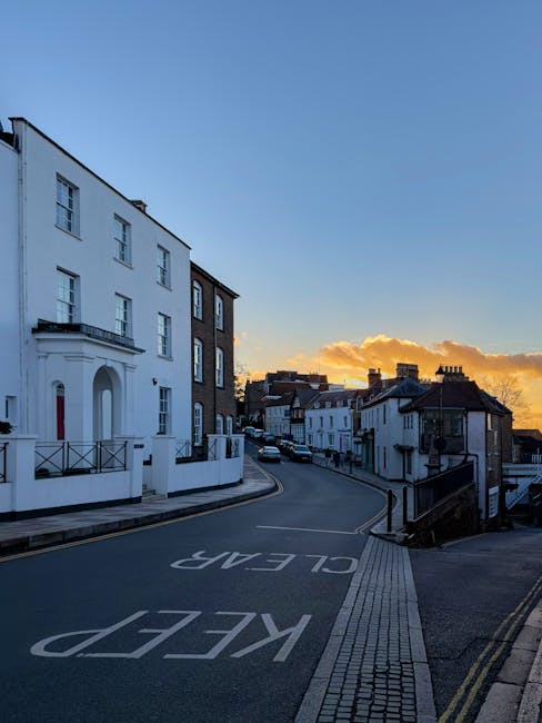 A quiet residential street at sunset, with a row of white and dark brick terraced houses lining the left side, and a gentle uphill slope leading to more buildings in the distance. The street surface is dark asphalt with clear white markings indicating a ‘KEEP CLEAR’ zone near the foreground, and a narrow pedestrian sidewalk with textured paving runs alongside. On the left, a large white building with a curved porch and decorative black railings stands prominently, while other houses further along have flat or gently pitched roofs, some with chimneys visible. The sky is brightly lit with warm orange and yellow hues from the setting sun, casting soft light and creating a serene atmosphere. The scene is free of clutter or rubbish, illustrating a well-maintained environment where rubbish clearance services such as those from Rubbish Clearance Harrow could support local property management and private waste handling solutions in Harrow on the Hill area, especially for residents requiring professional rubbish removal.