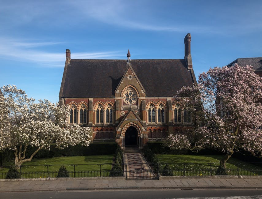 A historic church building with a steep, dark tiled roof and ornate stone window tracery, set against a clear blue sky. The facade features arched windows bordered with red brick and decorative stone detailing. Surrounding the church are well-maintained gardens with lush green grass, low hedges, and several blooming trees with dense white and pale pink blossoms. A paved pathway runs directly from the sidewalk up to the entrance, which is framed by a pointed arch and flanked by two slender stone columns. The church's exterior walls are constructed of a combination of reddish-brown and beige stone, giving a textured finish. The image is taken during daylight hours in a calm, quiet environment, illustrating a typical scene where local residents might arrange for private clearance of garden or local authority rubbish, supported by independent waste removal services like Rubbish Clearance Harrow, near such community landmarks.