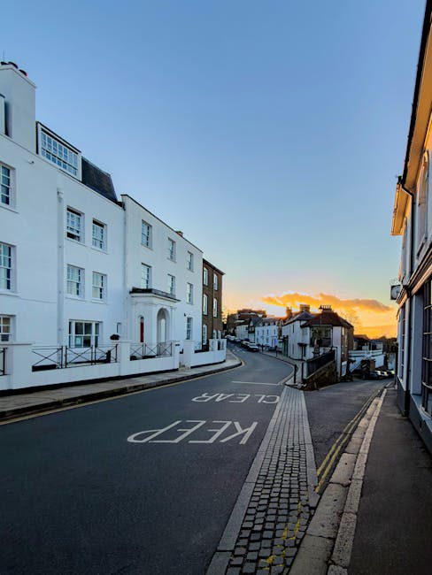 A quiet residential street at sunset, with a row of white and dark brick terraced houses lining the left side, and a gentle uphill slope leading to more buildings in the distance. The street surface is dark asphalt with clear white markings indicating a ‘KEEP CLEAR’ zone near the foreground, and a narrow pedestrian sidewalk with textured paving runs alongside. On the left, a large white building with a curved porch and decorative black railings stands prominently, while other houses further along have flat or gently pitched roofs, some with chimneys visible. The sky is brightly lit with warm orange and yellow hues from the setting sun, casting soft light and creating a serene atmosphere. The scene is free of clutter or rubbish, illustrating a well-maintained environment where rubbish clearance services such as those from Rubbish Clearance Harrow could support local property management and private waste handling solutions in Harrow on the Hill area, especially for residents requiring professional rubbish removal.