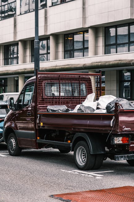 A small dark red flatbed truck parked in an urban environment with a modern building in the background featuring large glass windows and concrete panels. The truck's cargo bed is loaded with various types of waste materials, including black plastic rubbish bags, some partially open revealing mixed household waste. The vehicles are situated on a paved street with visible road markings, and the surrounding area includes other parked cars. The image scene depicts a typical private waste collection or rubbish removal service, with the truck prepared for on-site clearance of general waste, consistent with the kind of private rubbish disposal handled by companies such as Rubbish Clearance Harrow. Natural daylight illuminates the scene, highlighting the textures of the waste and the reflective surfaces of the vehicle. The setting suggests an area where independent rubbish collection services operate to facilitate timely waste removal, offering an alternative to local authority disposal channels.