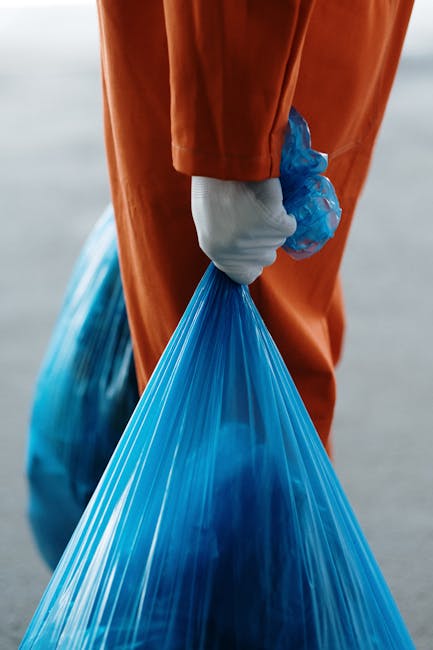 A close-up view of a person dressed in orange protective overalls holding a blue plastic rubbish bag, which is partially filled and sealed, with their hand encased in a white glove. The background suggests an outdoor environment, possibly a paved or concrete surface, with a blurred, neutral-toned setting. The focus is on the individual’s lower torso and arm, emphasizing the handling of waste in a careful, hygienic manner. The use of sturdy gloves and the plastic bag indicates proper handling of rubbish, aligning with waste management practices. This scene reflects procedures involved in private waste disposal or on-site rubbish clearance, often undertaken independently outside of local authority collections. The image underscores the importance of safety equipment during rubbish removal, which is a key aspect of professional rubbish clearance services like those provided by Rubbish Clearance Harrow, especially in contexts requiring urgent or specialized waste removal from rental properties or private premises.