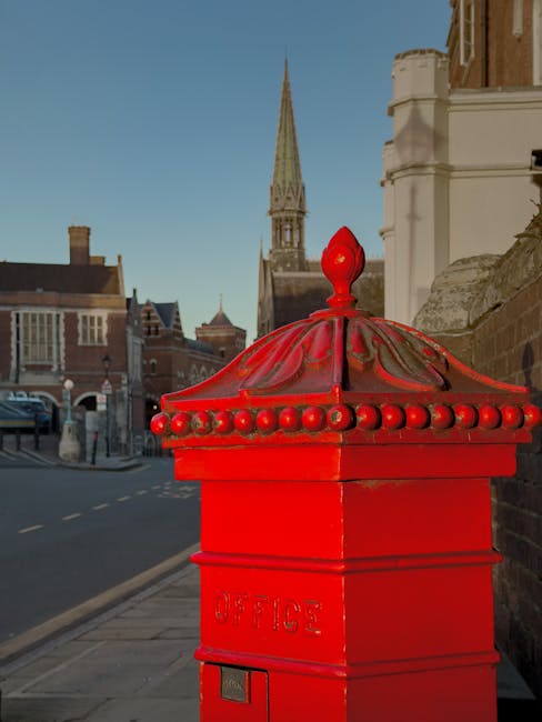 Close-up view of a bright red traditional British post box with a rounded, domed top adorned with decorative metal beads, situated on a pavement in an urban setting. Behind the post box, there are historic brick buildings with large sash windows and a tall, pointed church spire visible against a clear blue sky. The side of a white building with detailed architectural features is partially visible to the right, and a low brick wall runs along the street. The scene is illuminated by natural daylight, highlighting the vivid colour and metallic finish of the post box, which contrasts with the more muted tones of the surrounding buildings and street. The image captures a moment of everyday urban life, where the traditional postal infrastructure stands alongside historic architecture, exemplifying a typical central city street scene with visual relevance to the notions of local postal services or alternative collection points that connect to waste management and collection services provided by companies like Rubbish Clearance Harrow in private waste handling contexts.
